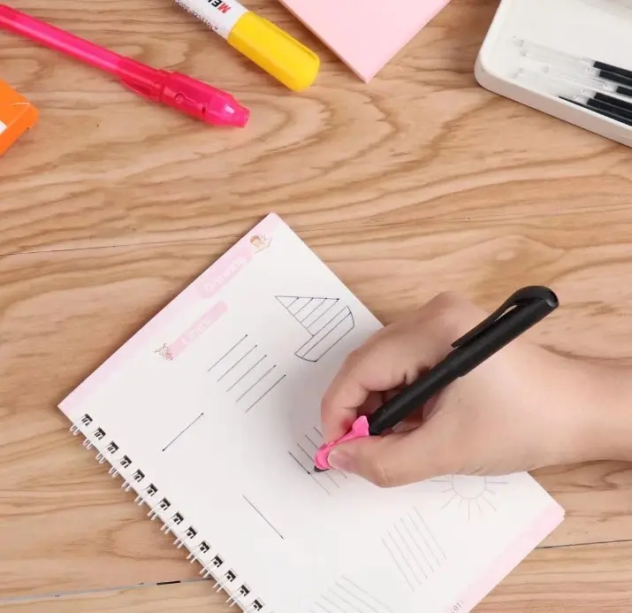 Mano escribiendo con bolígrafo negro y rosa en cuaderno de práctica mágica con espiral, líneas de caligrafía y bordes rosas sobre mesa de madera.