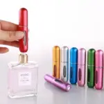 Atomizador de perfume portátil rojo recargable siendo llenado desde una botella, acompañado de varios frascos metálicos de colores en la superficie.