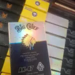 Caja de cartucho Big Chief AK-47 de 1000mg con acabado holográfico y fondo azul oscuro, exhibida en un estante de productos.