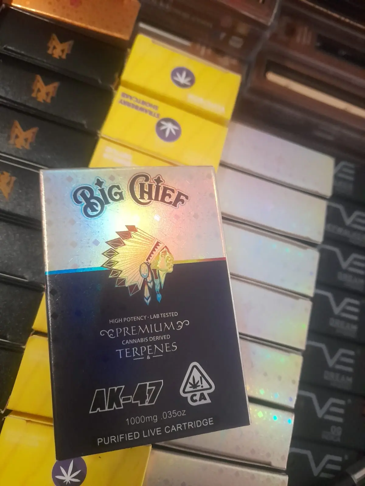 Caja de cartucho Big Chief AK-47 de 1000mg con acabado holográfico y fondo azul oscuro, exhibida en un estante de productos.