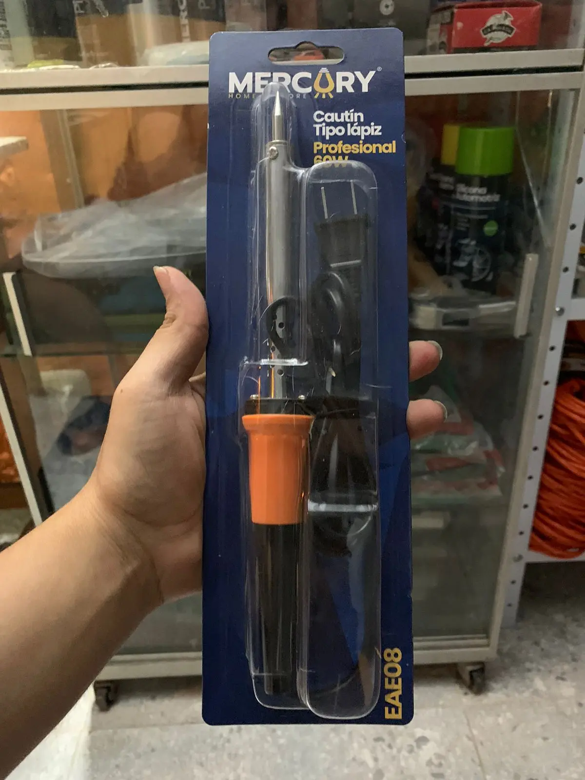 Mano sosteniendo cautín tipo lápiz de 60W con mango naranja y negro, empacado en blíster azul de la marca Mercury.