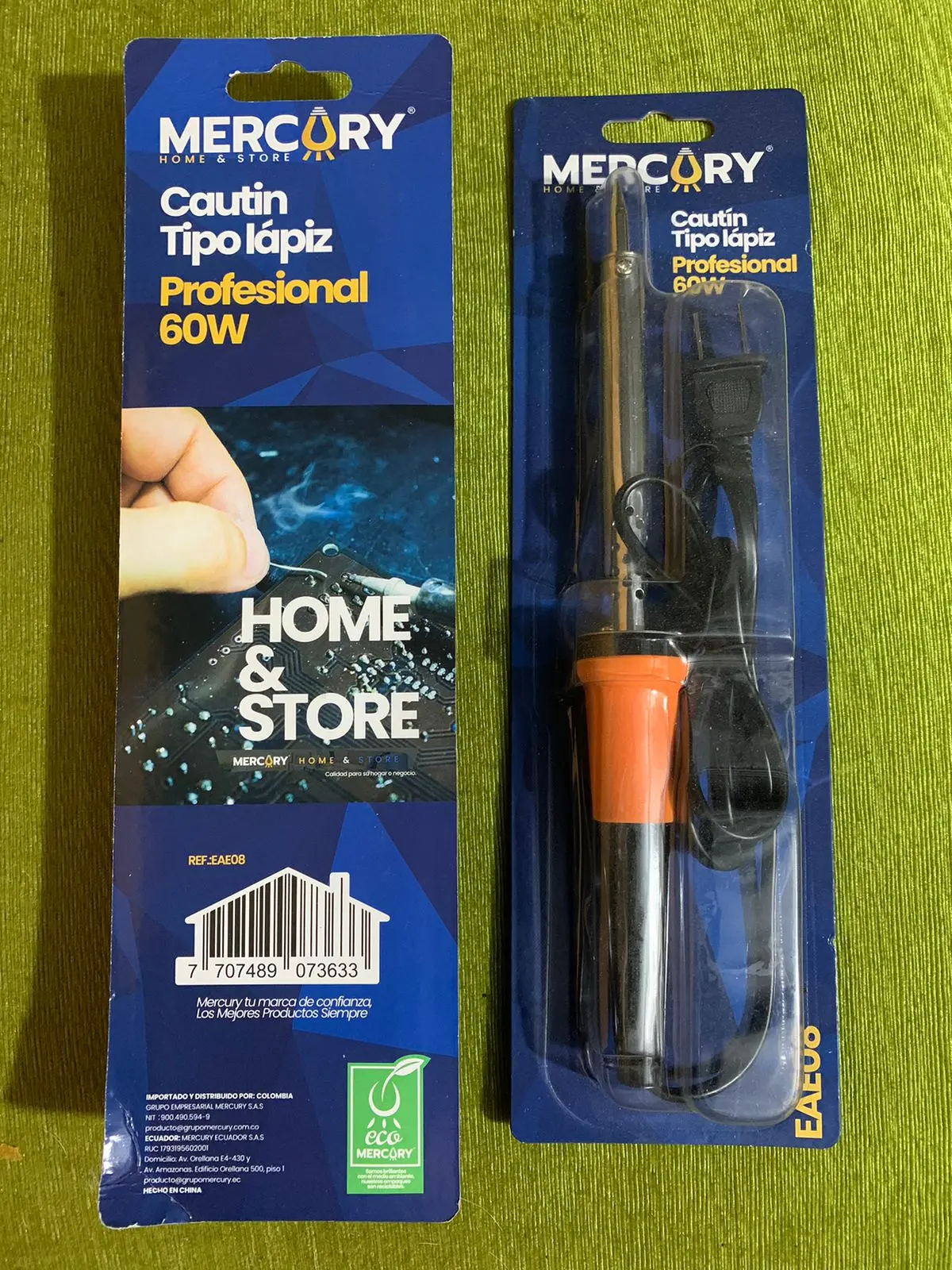 Cautín tipo lápiz Mercury de 60W con mango naranja y punta fina, en empaque de blíster con cable, junto a su cartón sobre fondo verde.