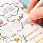 Mano usando marcador roller de punta doble color rosa para trazar ondas decorativas en cuaderno cuadriculado con dibujos kawaii.