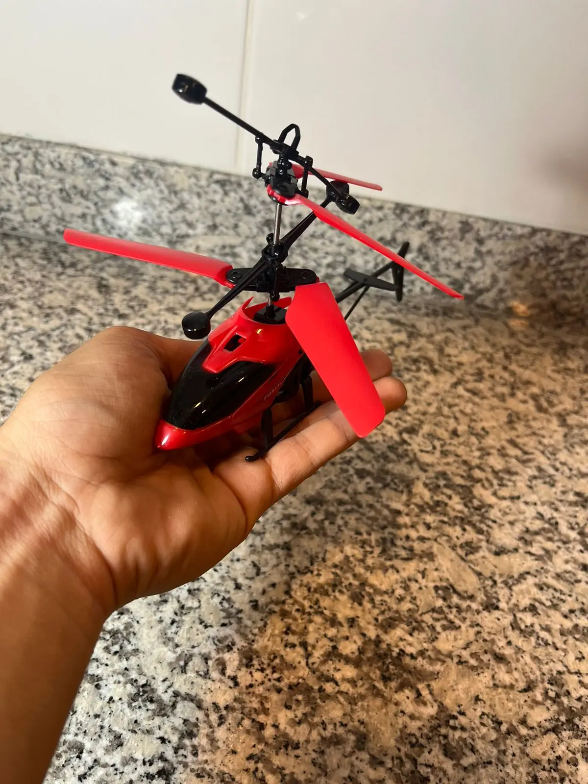 Helicóptero rojo a control remoto con sensor de movimiento y aspas flexibles, sostenido en la mano sobre fondo de granito.