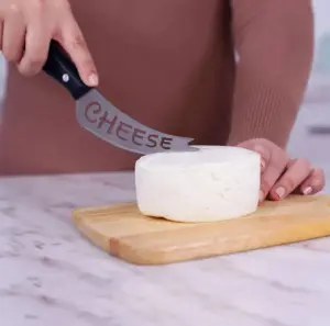 Cuchillo de acero inoxidable con mango negro y la palabra CHEESE grabada, cortando un queso blanco sobre una tabla de madera.