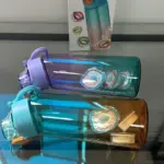 Dos botellas de agua saborizada degradadas con tapa de rosca y boquilla, una morada y otra turquesa con naranja, acompañadas de su caja blanca de empaque y anillos aromáticos en el interior.