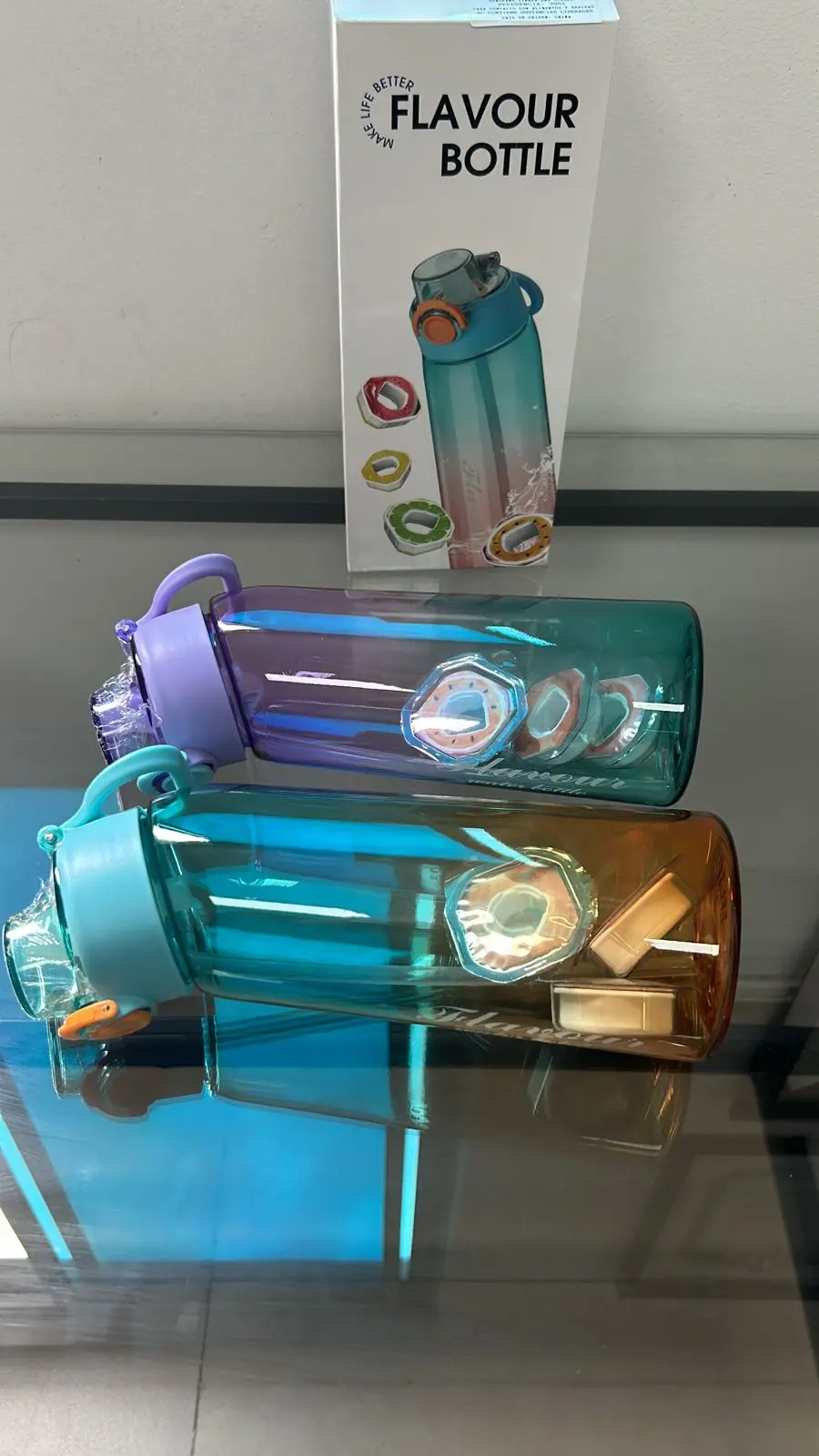 Dos botellas de agua saborizada degradadas con tapa de rosca y boquilla, una morada y otra turquesa con naranja, acompañadas de su caja blanca de empaque y anillos aromáticos en el interior.
