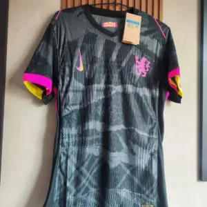 Camiseta de fútbol Chelsea tercera equipación en negro con estampado gris, detalles en fucsia y amarillo, colgada en percha.
