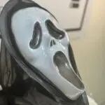 Máscara blanca de expresión de grito con capucha de tela negra tipo malla, vista lateral en primer plano para disfraz de Halloween.