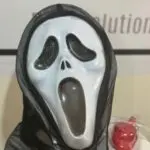 Máscara blanca de Scary Movie con boca abierta, capucha de tela negra translúcida y un accesorio rojo de utilería al lado.