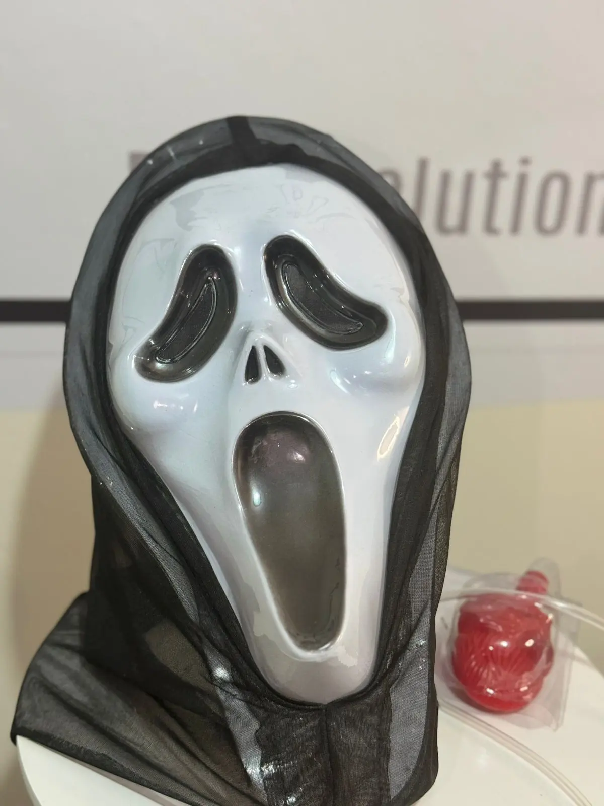 Máscara blanca de Scary Movie con boca abierta, capucha de tela negra translúcida y un accesorio rojo de utilería al lado.