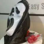 Máscara blanca de Halloween con capucha negra y bolsa de sangre artificial roja con tubo sobre superficie blanca.