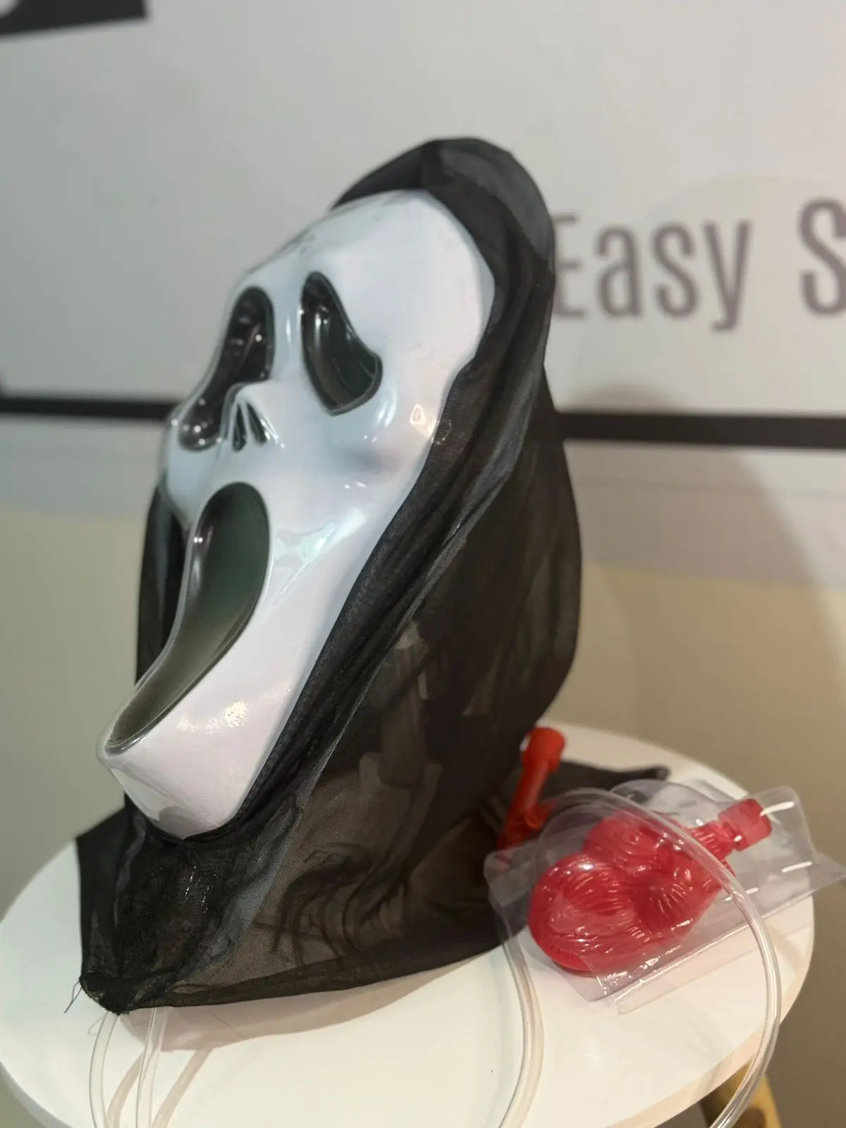 Máscara blanca de Halloween con capucha negra y bolsa de sangre artificial roja con tubo sobre superficie blanca.