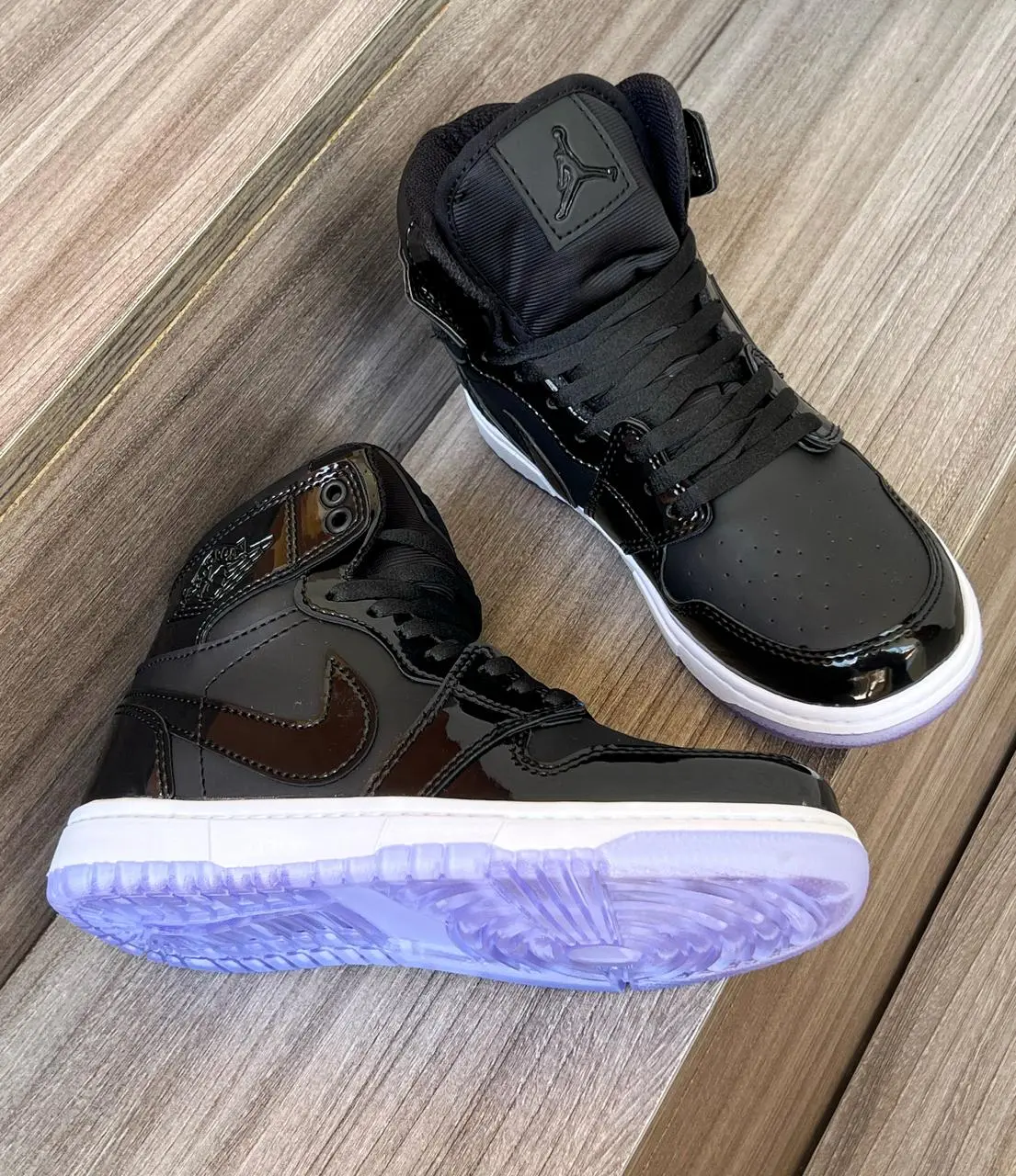 Tenis deportivos Jordan de caña alta en charol negro brillante con suela translúcida violeta, vistos desde ángulo superior y lateral sobre superficie de madera.