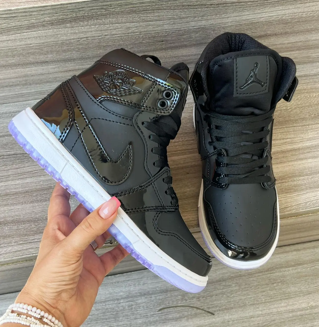 Tenis Jordan negros de caña alta en charol brillante con suela morada translúcida, sostenidos en mano sobre superficie de madera.