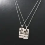 Par de collares de acero inoxidable con dijes rectangulares grabados con una pareja y frase dividida sobre fondo negro texturizado.
