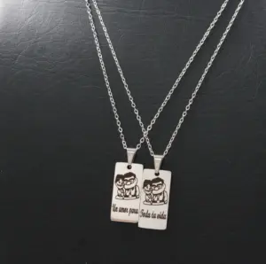 Par de collares de acero inoxidable con dijes rectangulares grabados con una pareja y frase dividida sobre fondo negro texturizado.