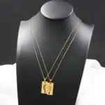 Collares de pareja dorados en acero inoxidable con dijes rectangulares de león y flecha, exhibidos sobre maniquí negro.