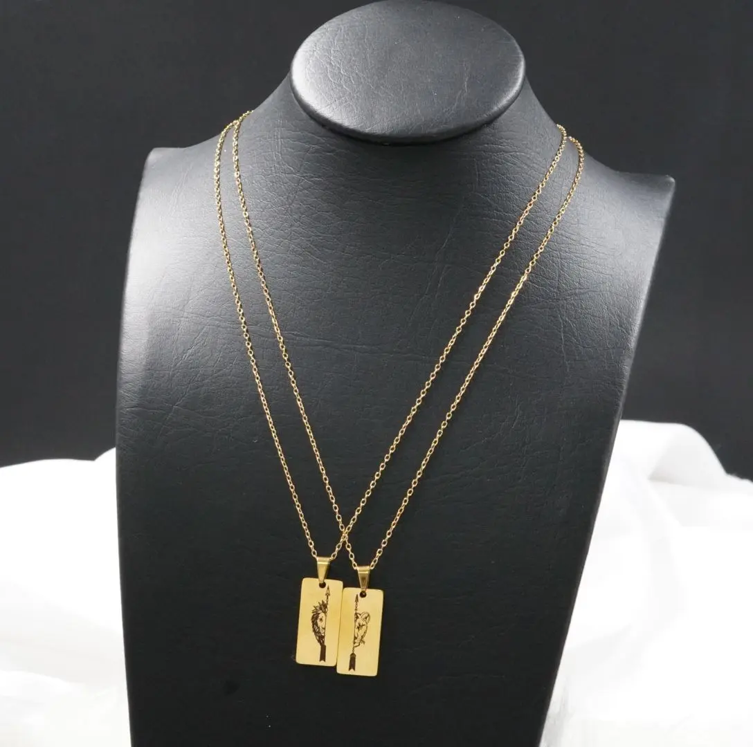 Collares de pareja dorados en acero inoxidable con dijes rectangulares de león y flecha, exhibidos sobre maniquí negro.