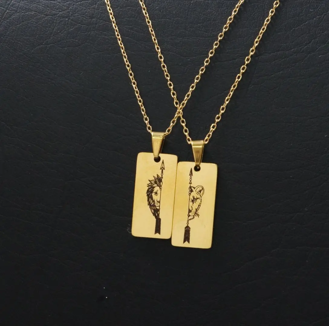 Dos collares dorados de acero inoxidable con placas rectangulares grabadas que muestran mitades de un león y una flecha, sobre fondo negro.