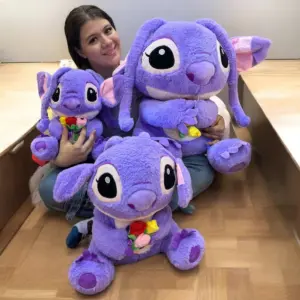 Peluche gigante morado Angel de 90 cm con flores en las manos, sentado en el piso junto a dos versiones más pequeñas.