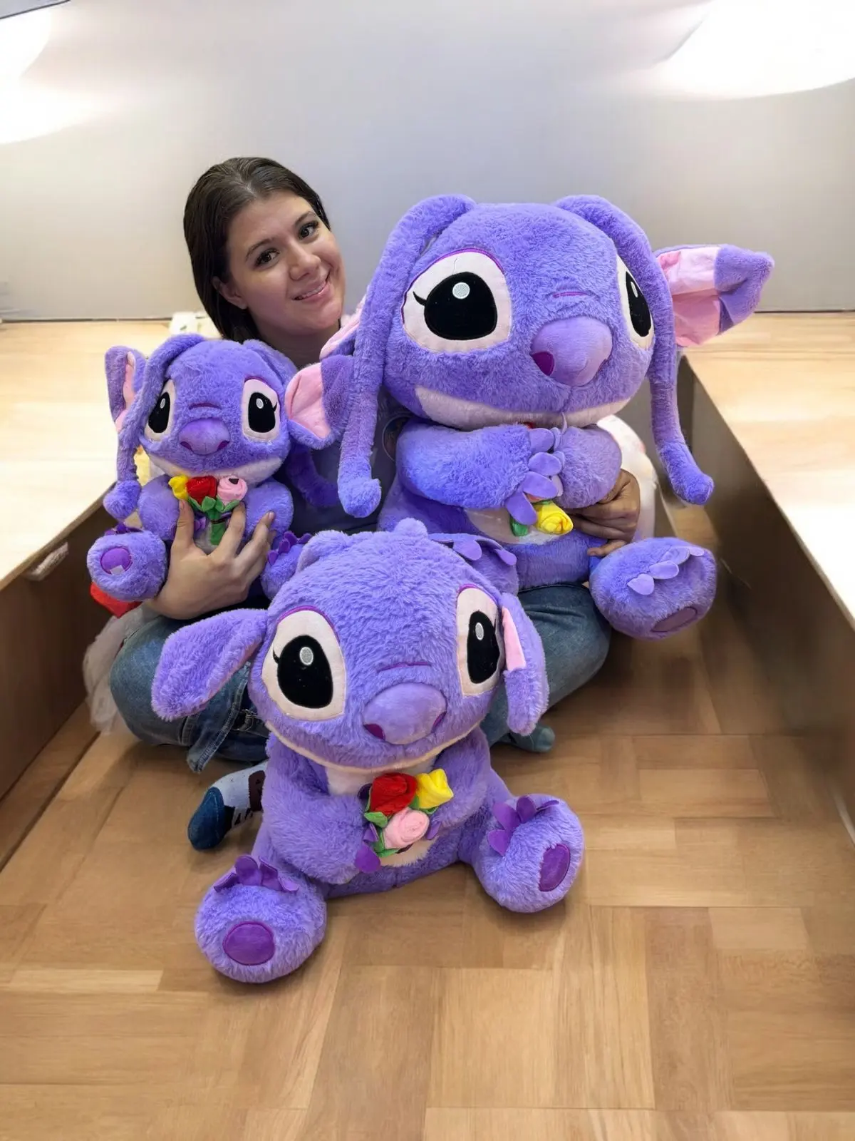 Peluche gigante morado Angel de 90 cm con flores en las manos, sentado en el piso junto a dos versiones más pequeñas.