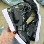 Tenis deportivos Jordan en charol negro de caña alta sostenidos en mano, con suela blanca y detalles brillantes.