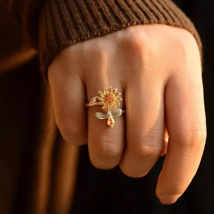 Anillo giratorio ajustable de girasol y abeja en metal dorado con circonitas, usado en el dedo anular sobre fondo oscuro.