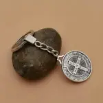 Llavero metálico plateado de San Benito con medalla grabada, unido a una argolla por cadena corta y presentado sobre una piedra gris.