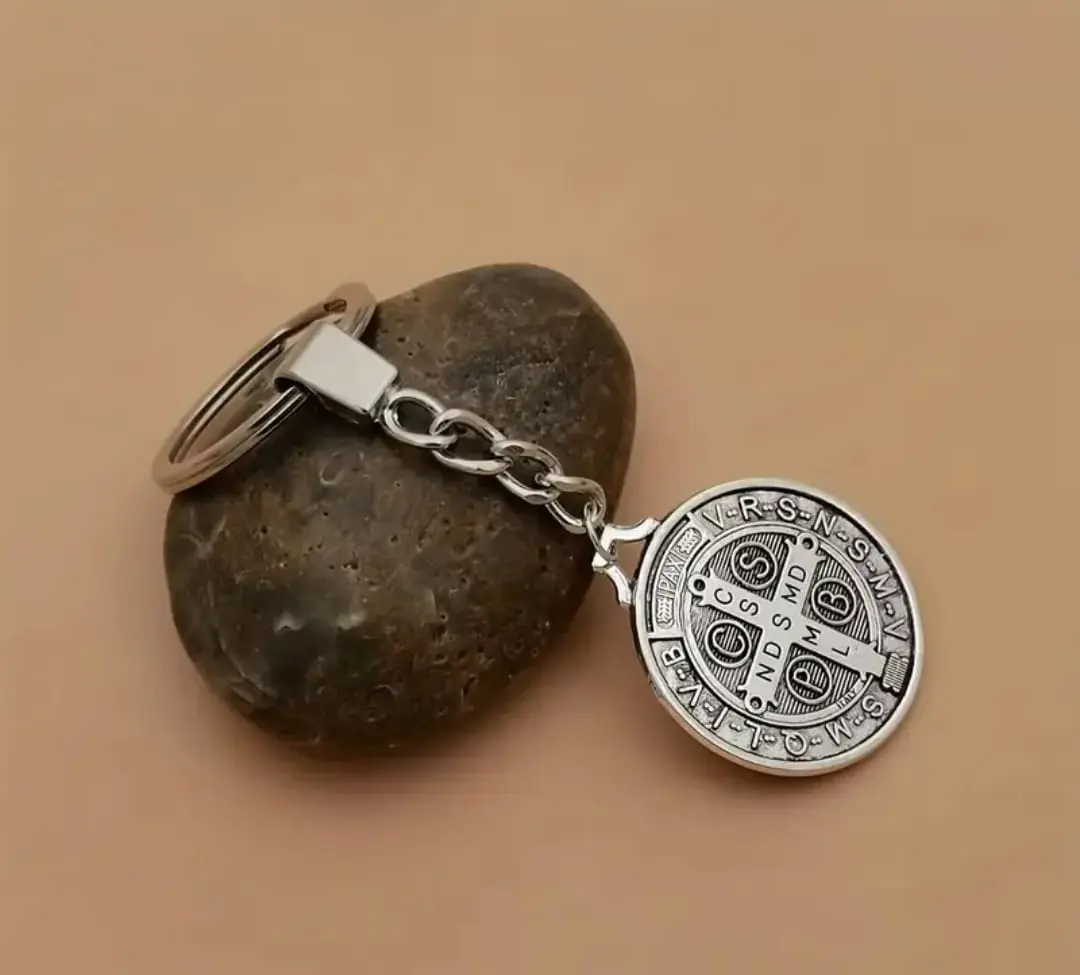 Llavero metálico plateado de San Benito con medalla grabada, unido a una argolla por cadena corta y presentado sobre una piedra gris.