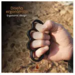 Manopla de acero negro con diseño ergonómico sujeta por una mano en puño para defensa personal, con fondo de tierra desenfocado.