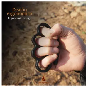 Manopla de acero negro con diseño ergonómico sujeta por una mano en puño para defensa personal, con fondo de tierra desenfocado.