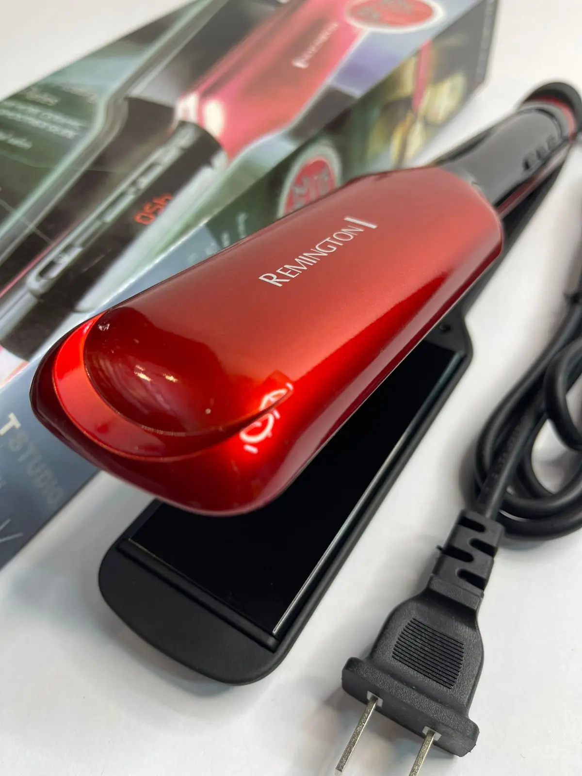 Plancha de cabello Remington roja con acabado brillante y placas anchas, apoyada junto a su caja y cable de alimentación.