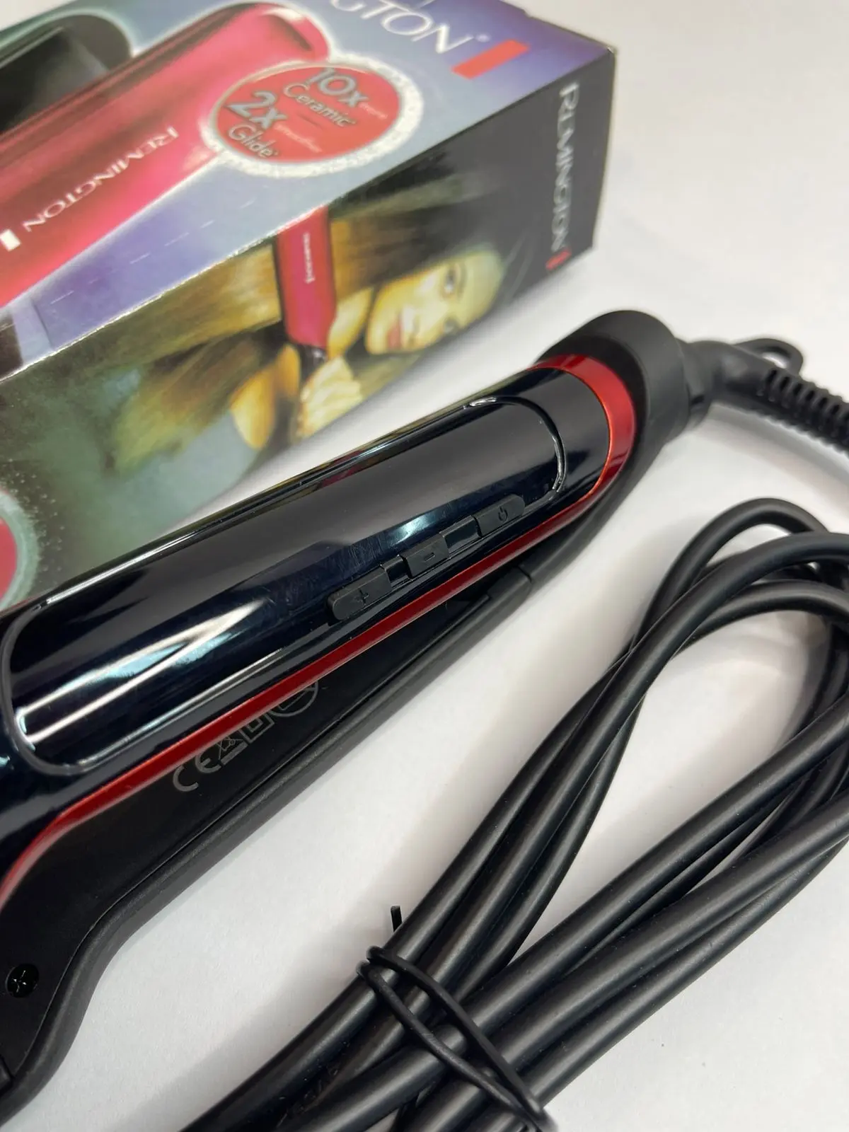 Plancha de cabello Remington roja con placas anchas de microcerámica, cable negro enrollado y caja original al fondo.