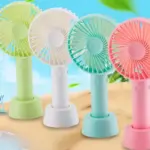 Cuatro ventiladores portátiles de mano en colores verde, blanco, celeste y rosado, con base redonda y aspas visibles sobre arena de playa.