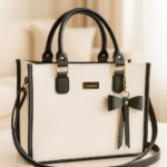 Bolso estructurado de cuero sintético blanco con detalles negros, herrajes dorados, placa con la marca Tahima, lazo decorativo colgante y correa larga ajustable.