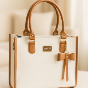 Bolso de mano estructurado en cuerina color marfil con ribetes y asas marrones, detalles dorados, placa frontal y llavero con lazo decorativo sobre mesa.