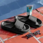 Par de sandalias negras antideslizantes de suela EVA texturizada, junto a gafas de sol y protector solar sobre borde de piscina.