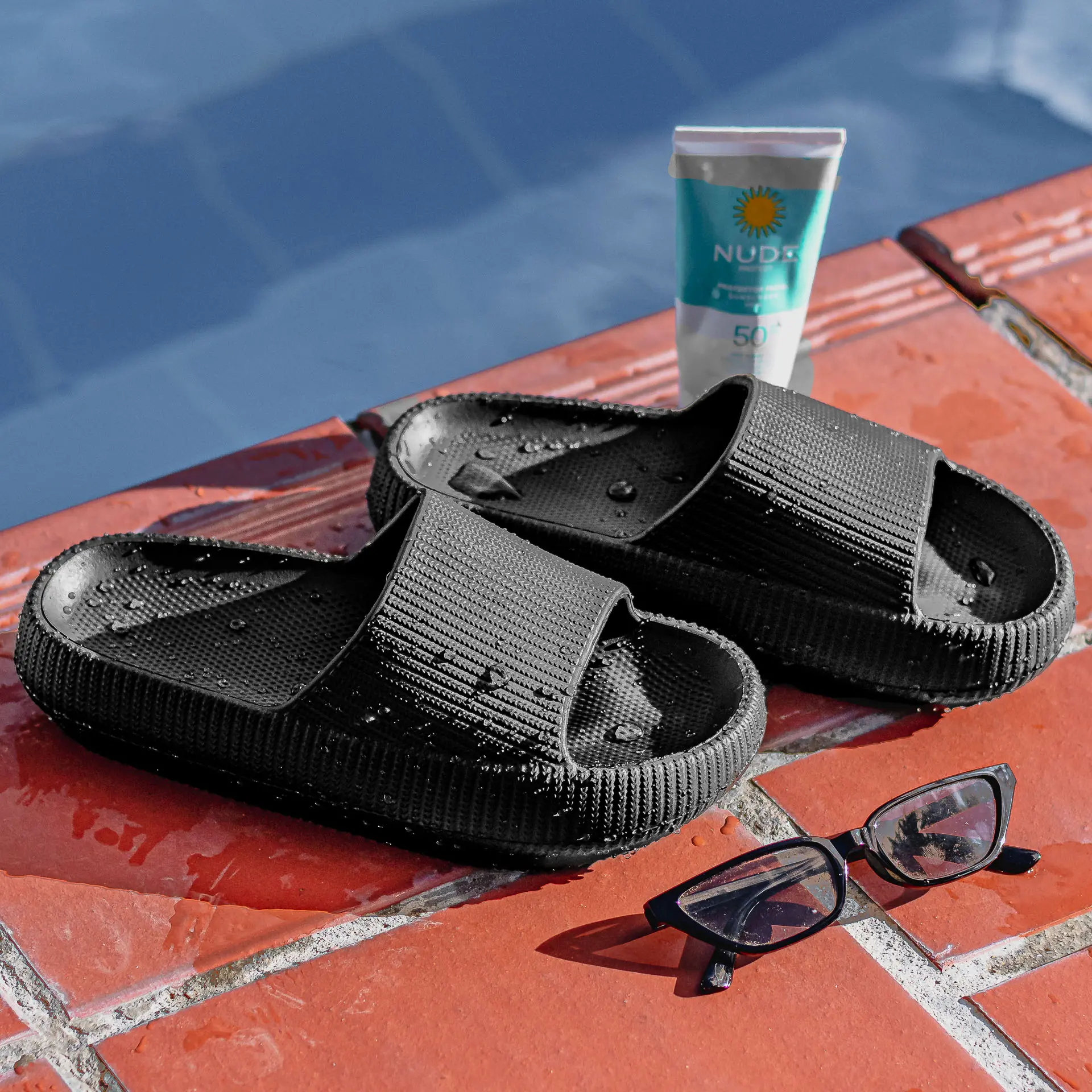 Par de sandalias negras antideslizantes de suela EVA texturizada, junto a gafas de sol y protector solar sobre borde de piscina.
