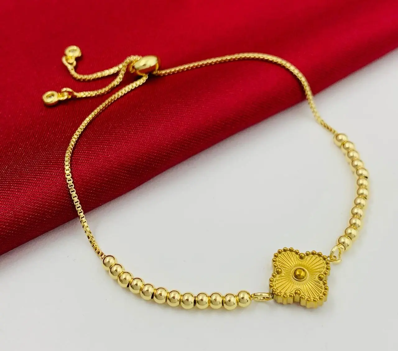 Pulsera dorada ajustable con abalorios redondos y dije central tipo flor estriada, dispuesta sobre tela roja y superficie blanca.