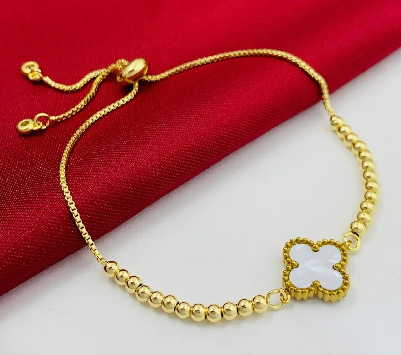 Pulsera dorada con cuentas tipo balín y dije central de trébol blanco, con cierre ajustable de cadena fina.