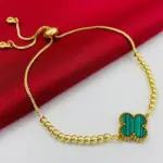 Pulsera dorada ajustable con dije de trébol verde rayado y cuentas esféricas sobre fondo rojo.