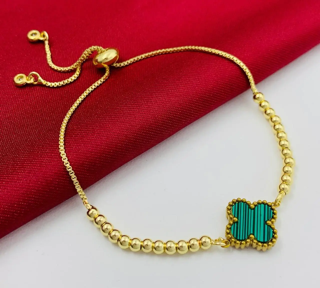 Pulsera dorada ajustable con dije de trébol verde rayado y cuentas esféricas sobre fondo rojo.