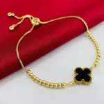 Pulsera dorada con dijes de balines, trébol negro central tipo Van Cleef y cierre ajustable deslizante sobre fondo rojo y blanco.
