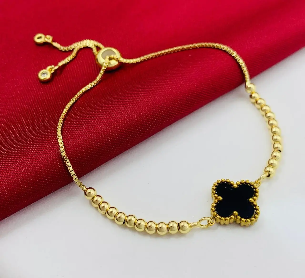 Pulsera dorada con dijes de balines, trébol negro central tipo Van Cleef y cierre ajustable deslizante sobre fondo rojo y blanco.