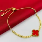 Pulsera dorada con cuentas tipo balín y dije de trébol rojo, con cierre ajustable, sobre fondo de tela roja y superficie blanca.