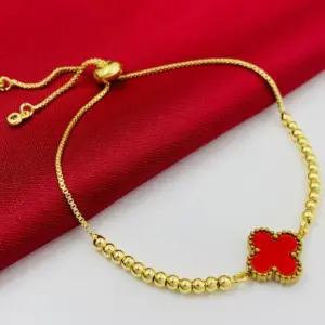 Pulsera dorada con cuentas tipo balín y dije de trébol rojo, con cierre ajustable, sobre fondo de tela roja y superficie blanca.