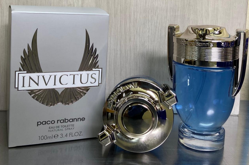 Perfume Paco Rabanne Invictus Eau de Toilette de 100 ml con frasco en forma de trofeo azul y plateado junto a su caja gris y alas.