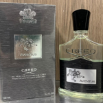 Perfume Creed Aventus de 100 ml con frasco de vidrio texturizado negro, líquido transparente y caja gris plomada con relieve plateado.