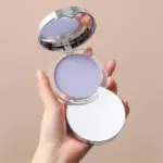 Polvo matificante compacto color lavanda en estuche transparente con espejos, sostenido por una mano.